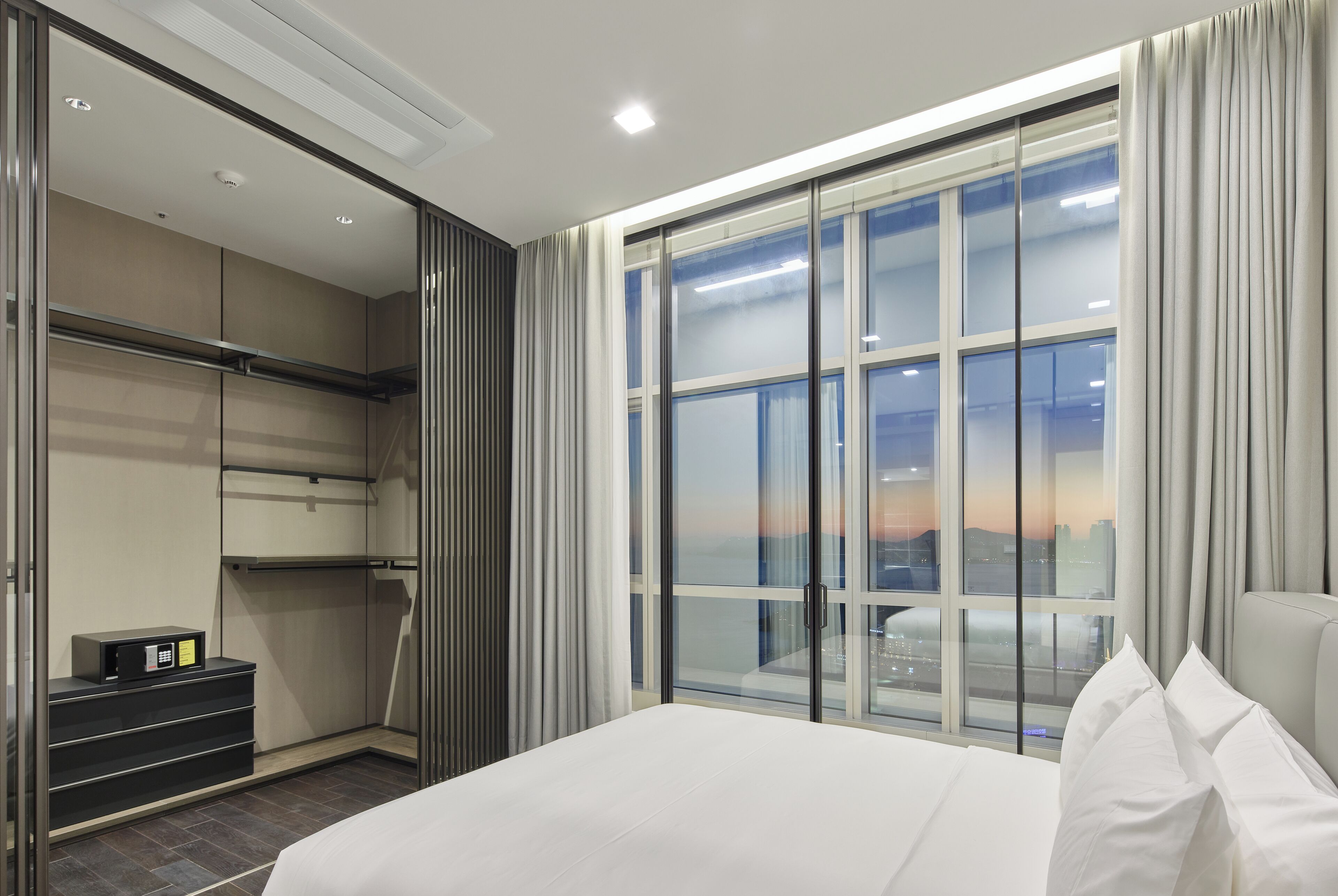 Ein modernes Hotelzimmer mit einem großen Bett, einem begehbaren Kleiderschrank und Blick auf die Stadt durch raumhohe Fenster.