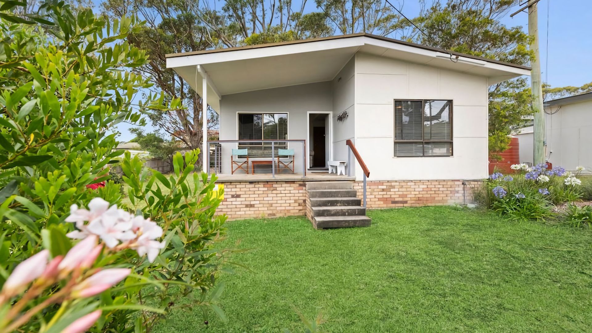 Uma casa moderna de dois andares com varanda coberta, base de tijolos e pequeno jardim frontal com grama e flores.