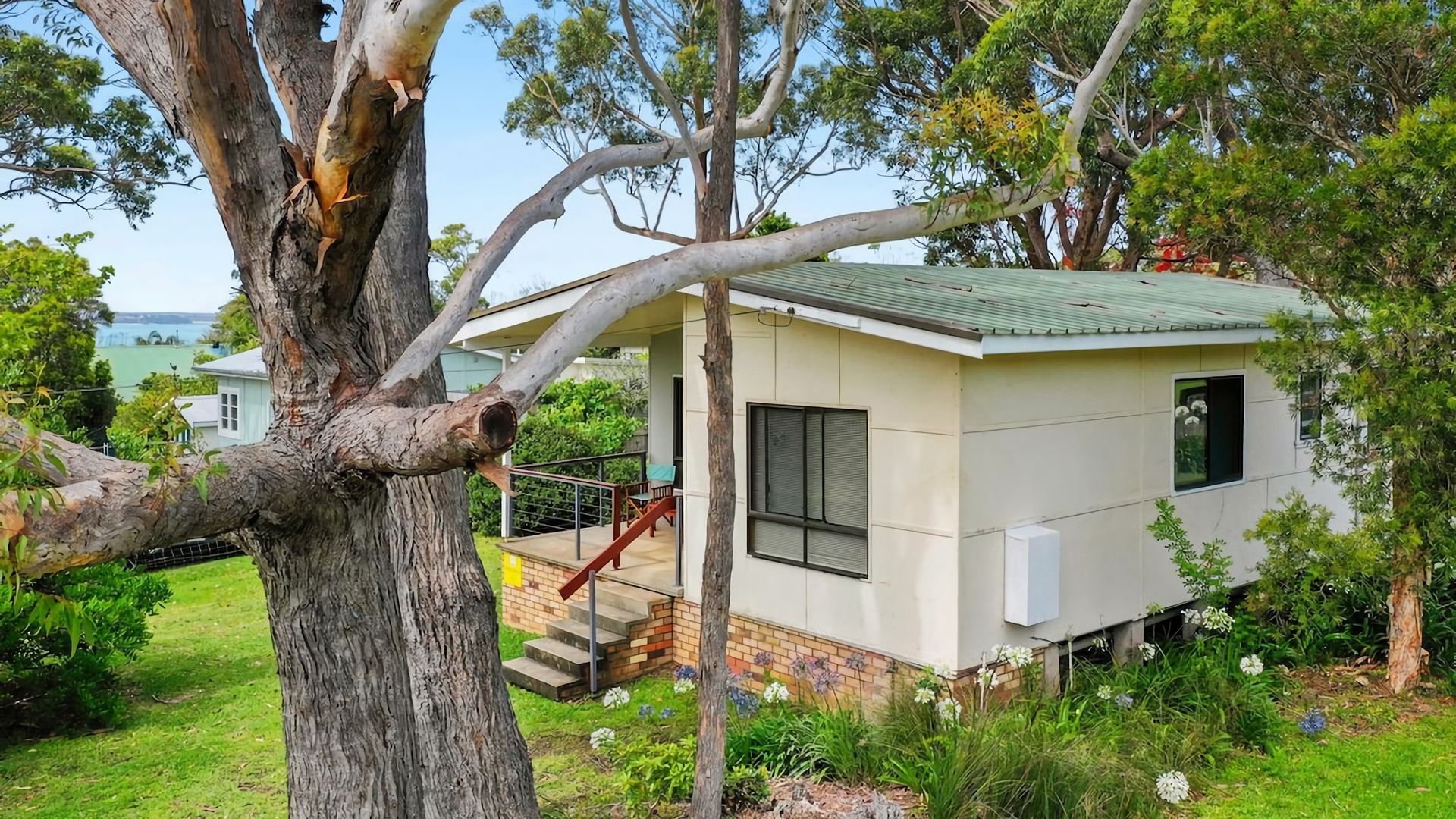 A small, single-story building with a balcony, surrounded by trees and a grassy area.