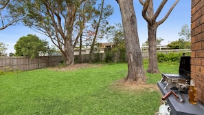 A backyard with a grassy lawn, a wooden fence, and a barbecue grill with utensils. - Kia-Ora near Washerwomans Beach (Bendalong)