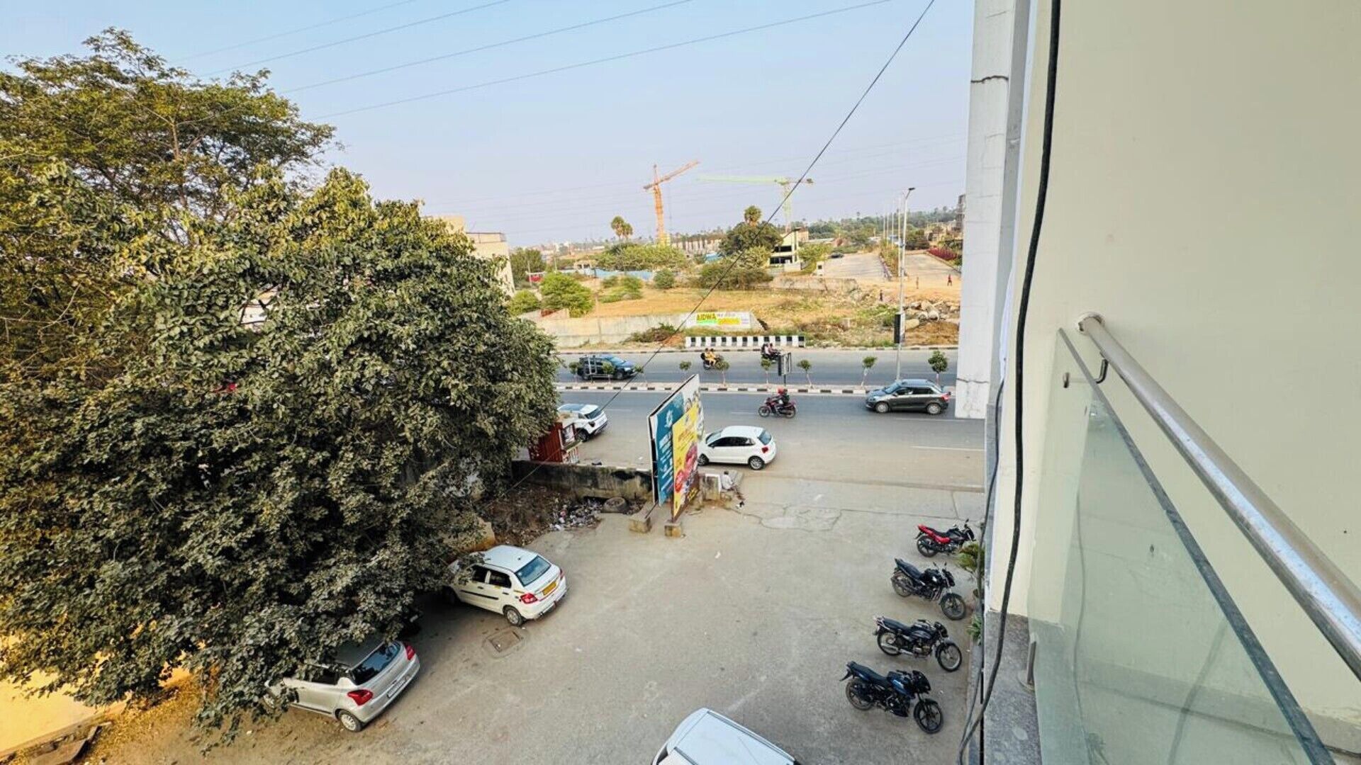 A view from a balcony overlooking a street with parked cars and motorcycles, a construction crane in the distance, and a large tree on the left.