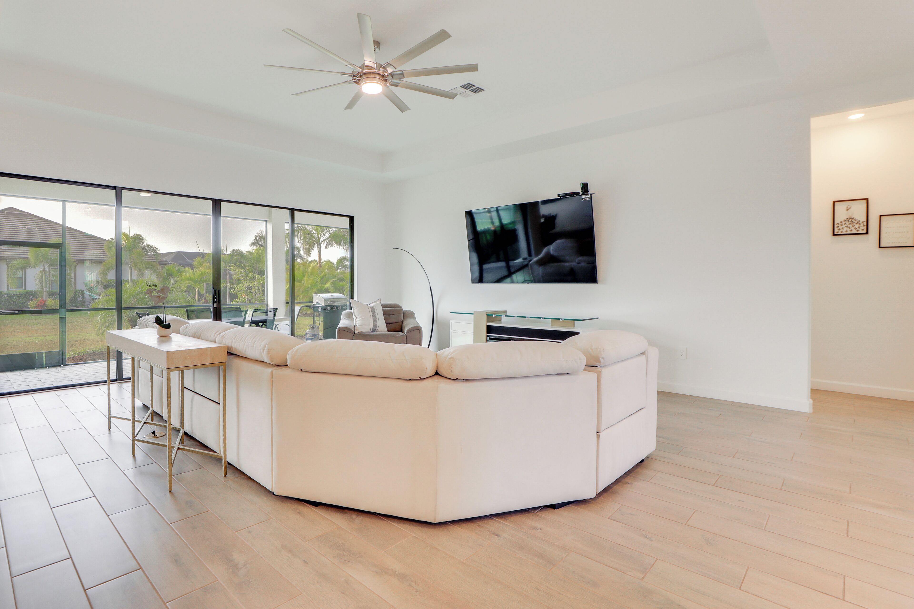 A modern living room with a large sectional sofa, a glass coffee table, a ceiling fan, and a TV mounted on the wall.
