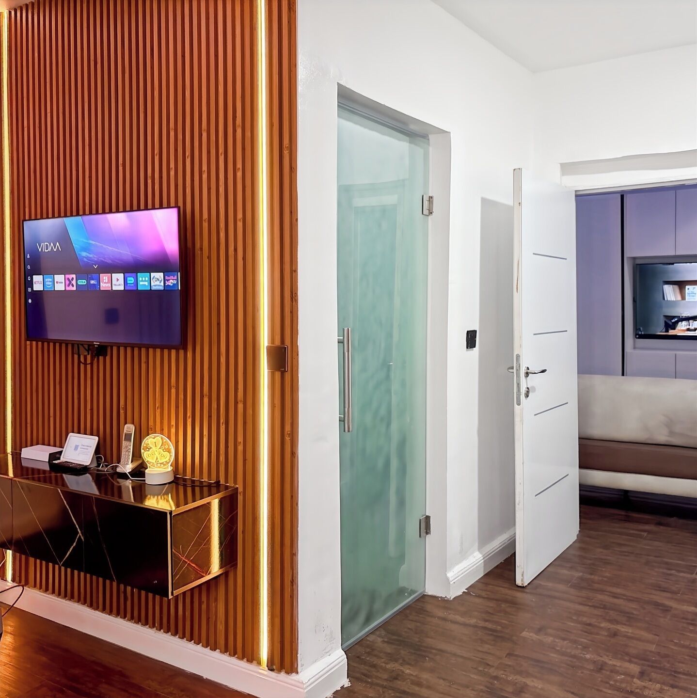 Ein modernes Wohnzimmer mit Flachbildfernseher an einer Holzwand, einer Glastür und einer Sitzecke mit Sofa und TV-Regal.