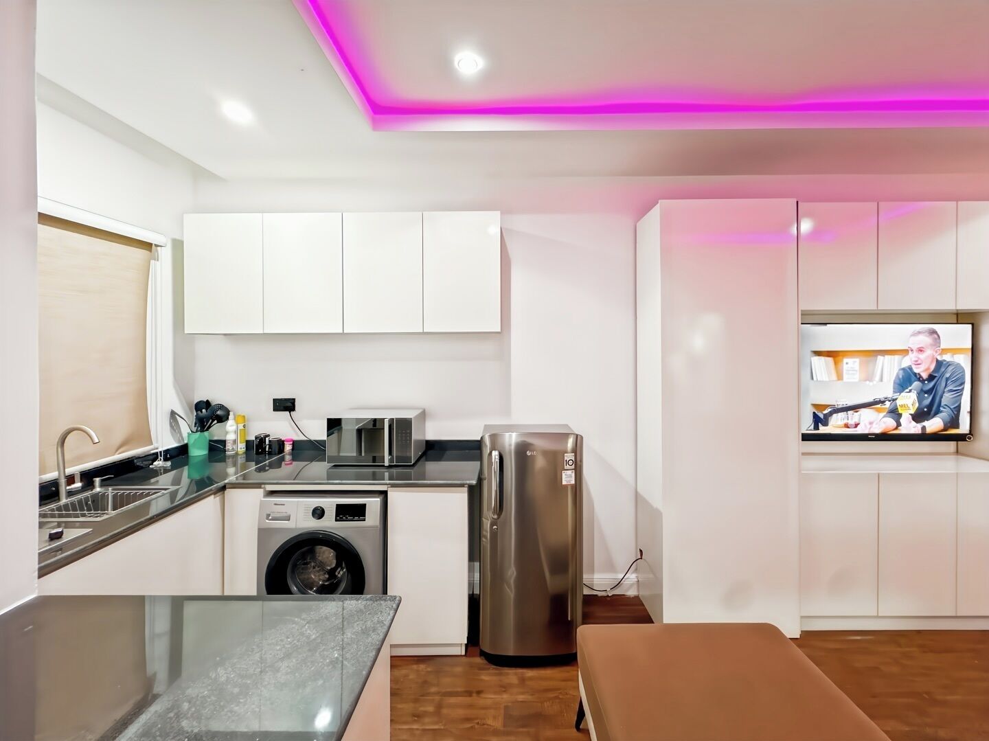 A compact kitchen with white cabinets, a washing machine, a refrigerator, a microwave, and a TV mounted on the wall.