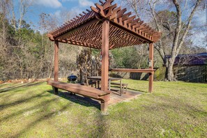 Ein hölzerner Pavillon mit Bänken und Tischen, umgeben von Bäumen und einer Rasenfläche.