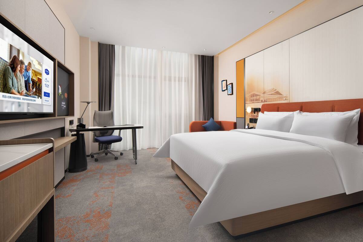 A hotel room with a large bed, a desk, a chair, and a TV screen displaying a group of people.