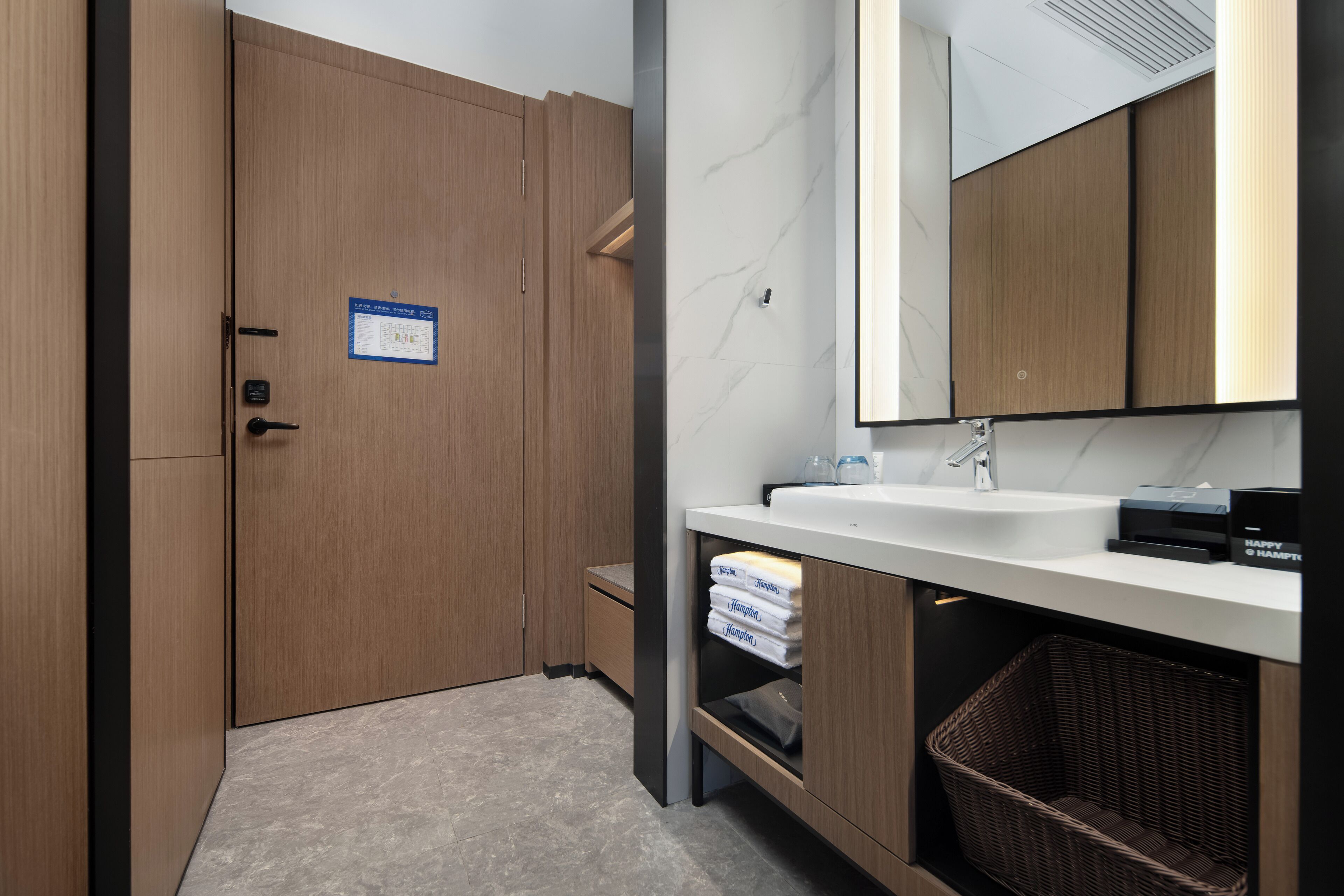 Un baño moderno con tocador de madera, un lavamanos y un espejo. Hay un cartel en la puerta, una cesta debajo del tocador y un cubo de basura.