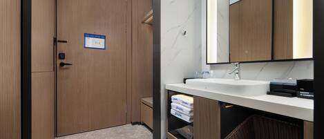 Un baño moderno con tocador de madera, un lavamanos y un espejo. Hay un cartel en la puerta, una cesta debajo del tocador y un cubo de basura.