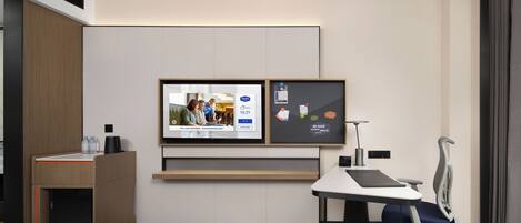 A hotel room with a bed, a desk with a chair, a TV mounted on the wall, and a small cabinet.