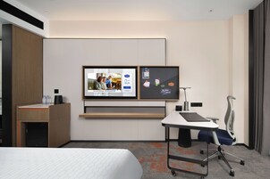 Habitación de hotel con una cama, un escritorio con silla, un televisor montado en la pared y un mueble auxiliar pequeño.