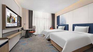 A hotel room with two beds, a desk, a chair, a TV, and a large window with curtains.