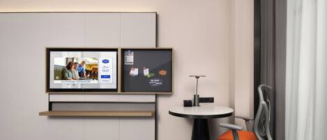 A hotel room with a bed, a TV mounted on the wall, a small round table with a chair, and a desk with a chair.