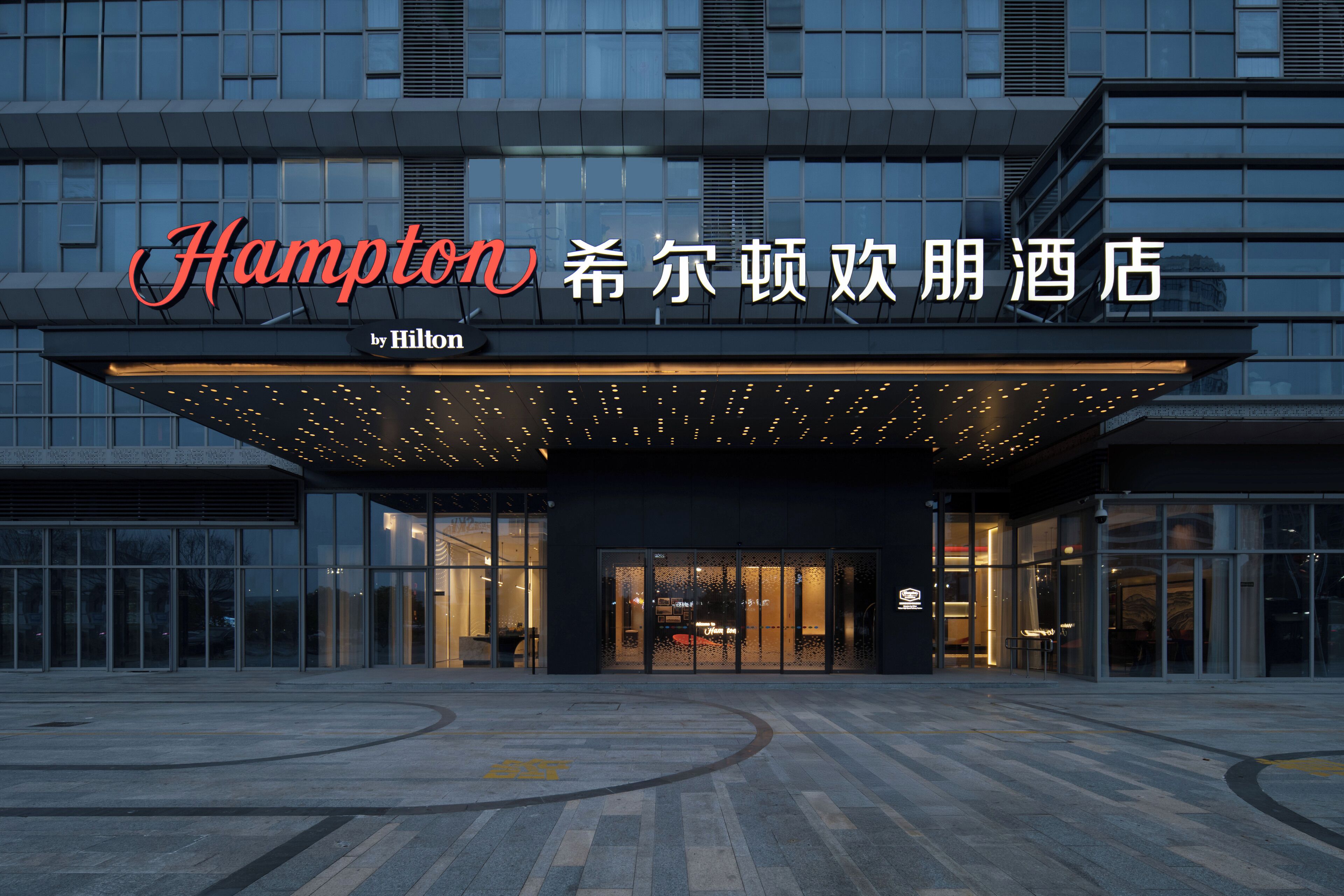 A modern hotel entrance with a large sign displaying the name "Hampton" and "Hilton" branding.