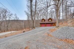 A two-story log cabin with a red garage door, surrounded by leaf-covered ground and trees. - Explore Nature & History! Getaway Near Gettysburg (Orrtanna)