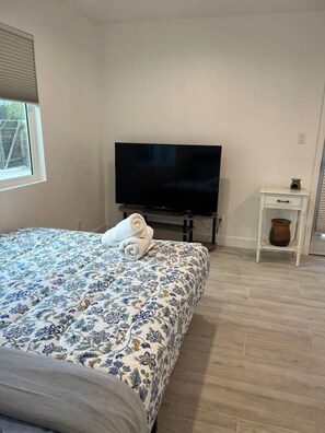 Ein Schlafzimmer mit einem Bett, einem Fernseher, einem kleinen weißen Nachttisch und einem Fenster.