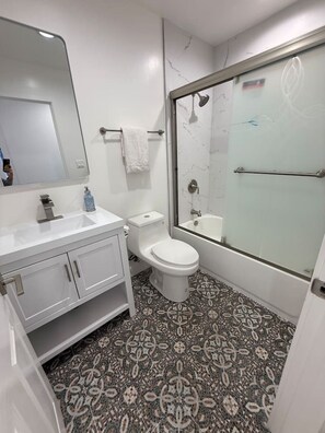 A modern bathroom with a white vanity, a toilet, a shower with a glass door, and patterned floor tiles. - Brand-New Home. Great Location. Monthly Discounts (Los Angeles)