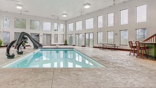 A spacious indoor pool area with a diving board, large windows, and a seating area with chairs and a table.