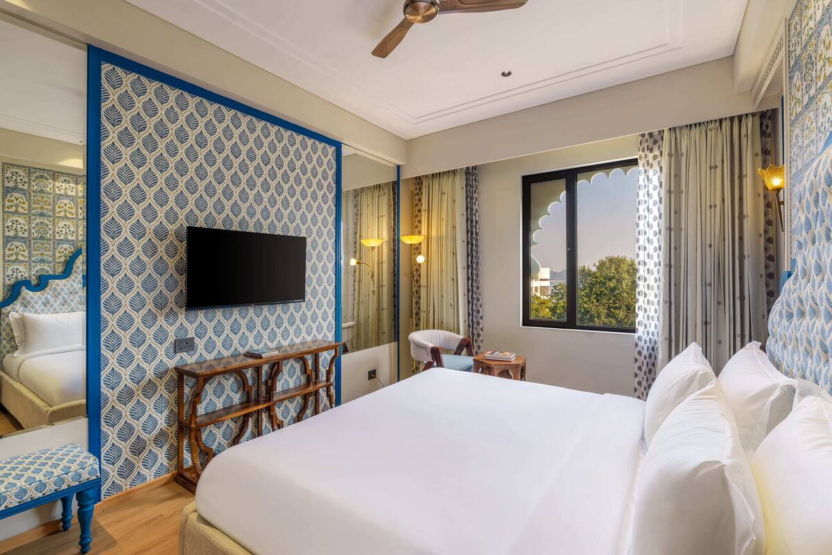 A hotel room with a large bed, a television mounted on the wall, a chair, a small table, and a window with curtains.