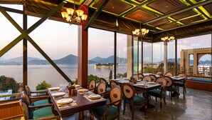 Un restaurante con terraza que da a un lago y montañas, con mesas y sillas de madera y ventanas grandes.