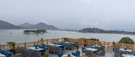 Eine Terrasse mit Korbmöbeln und blauen Kissen mit Blick auf einen stillen See, auf dem Boote liegen, und Hügel in der Ferne.