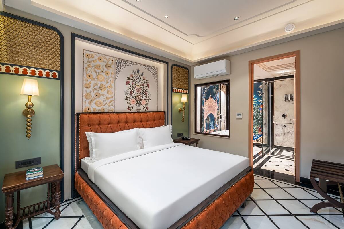 A hotel room with a large bed, a nightstand, a wall-mounted lamp, and a door leading to a bathroom.
