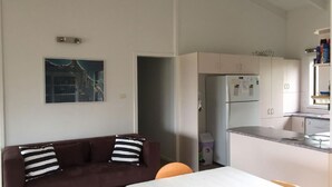 Un saló amb un sofà marró i almoadines a ratlles, una cuina amb armaris blancs i un frigorífic, i un quadre a la paret.
