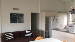 Un salon avec un canapé marron et des coussins à rayures, une cuisine avec des meubles blancs et un réfrigérateur, et un tableau accroché au mur.