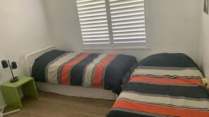 A bedroom with two beds, a nightstand, a lamp, and a window with blinds. - Life's A Beach with views to Green Island (Manyana)