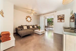 Ein modernes Wohnzimmer mit einer Couch, einem Couchtisch und einem Fernseher. Es gibt einen Deckenventilator, eine Wanduhr und ein Fenster mit Vorhängen.