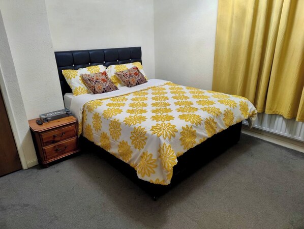 A neatly made bed with a floral-patterned bedspread, a wooden nightstand with a remote control, and a yellow curtain. - Peterborough Nice House With Garden Free Parking (Peterborough)