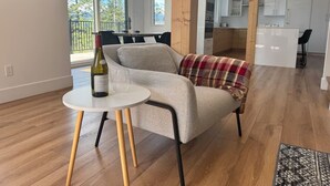 Ein modernes Wohnzimmer mit einer Couch, einem kleinen runden Tisch und einer Weinflasche. Große Fenster bieten einen Blick ins Freie.