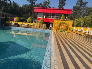 A swimming pool with a tiled edge, a wooden deck, and a building with a red roof in the background. - Forest king Resorts TDG Group (Rishikesh)
