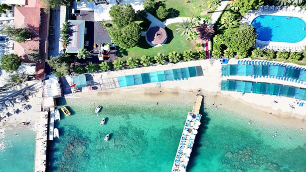 Vista aérea de un complejo turístico en la playa, con aguas turquesas cristalinas, un muelle largo con sombrillas y una zona de piscina con sillones de descanso.