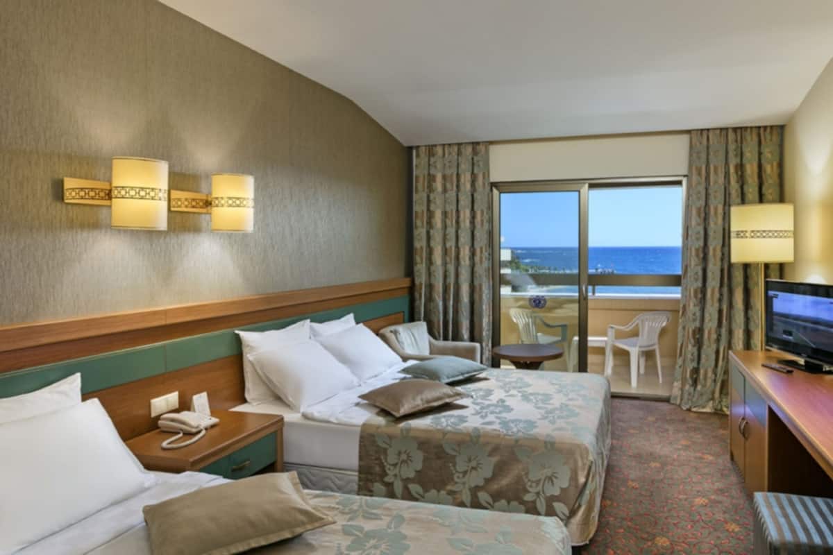 A hotel room with two beds, a TV, a desk, and a balcony with a view of the ocean.