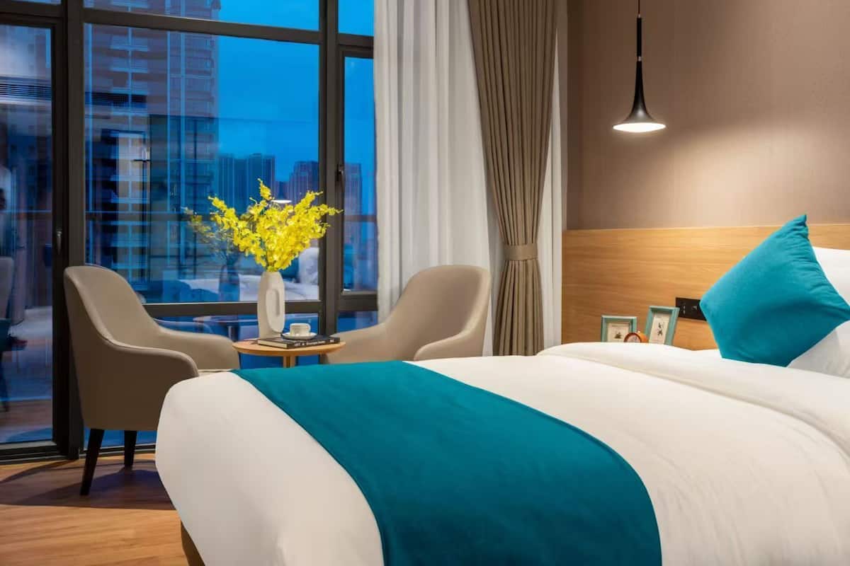 Ett hotellrum med en stor säng, två fåtöljer, en vas med gula blommor, utsikt över staden genom stora fönster och en modern hänglampa.