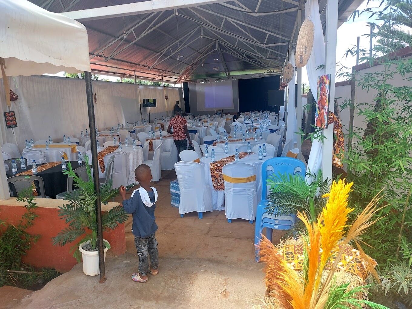 An indoor event space with tables set up for an event, surrounded by potted plants and a stage area.