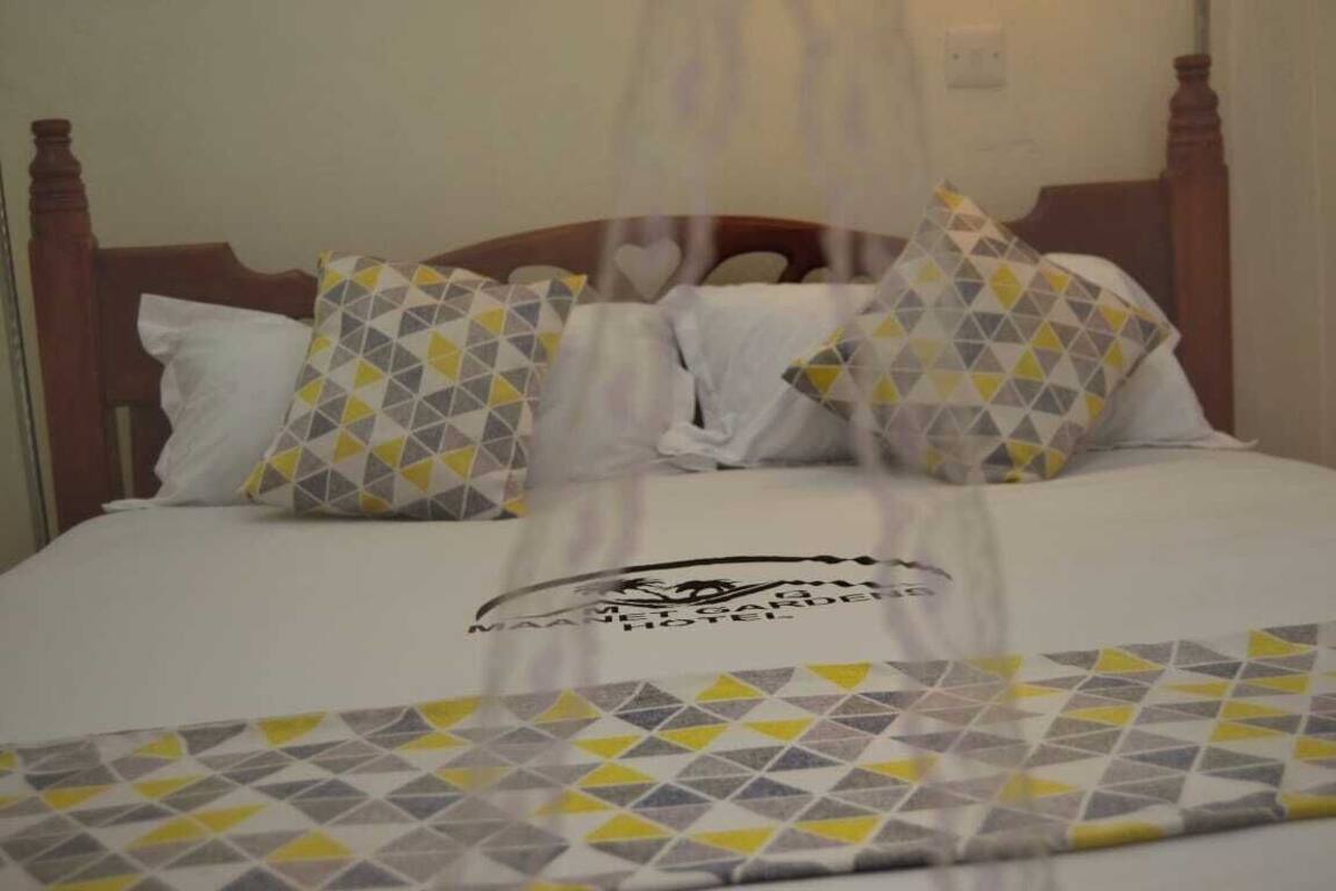 A bed with a geometric patterned bedspread and pillows, a wooden headboard, and a logo for the Maans Garden Hotel.