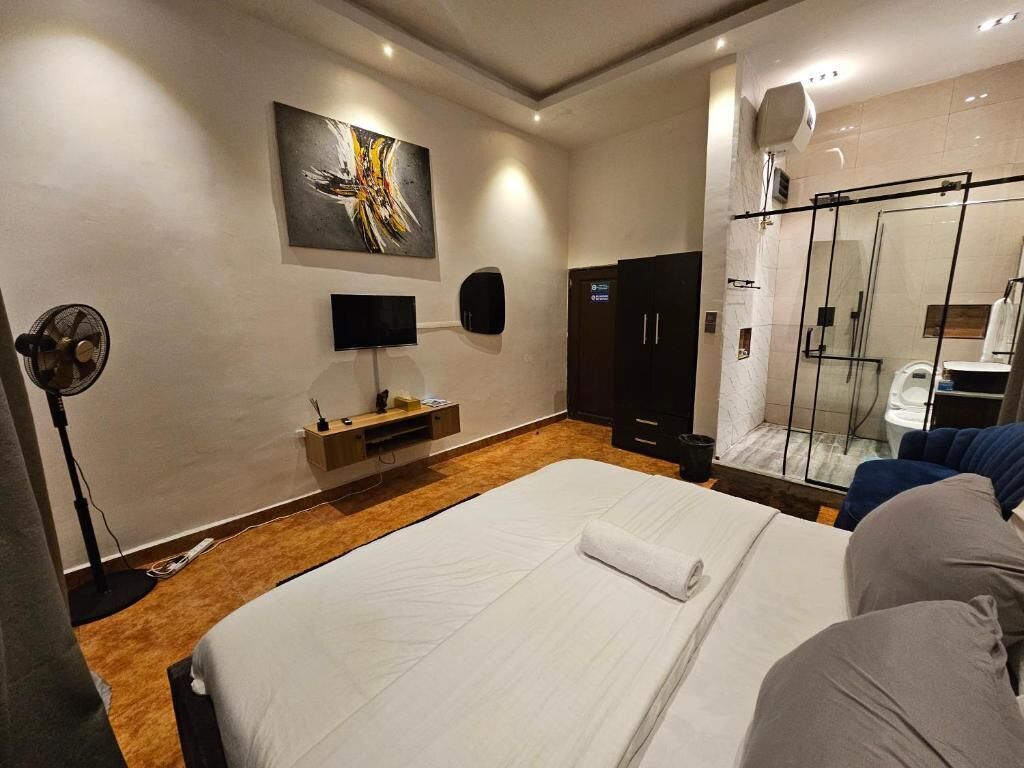 Moderni hotellihuone, jossa on suuri sänky, televisio, taulu, vaatekaappi ja lasiseinällä erotettu suihkutila.