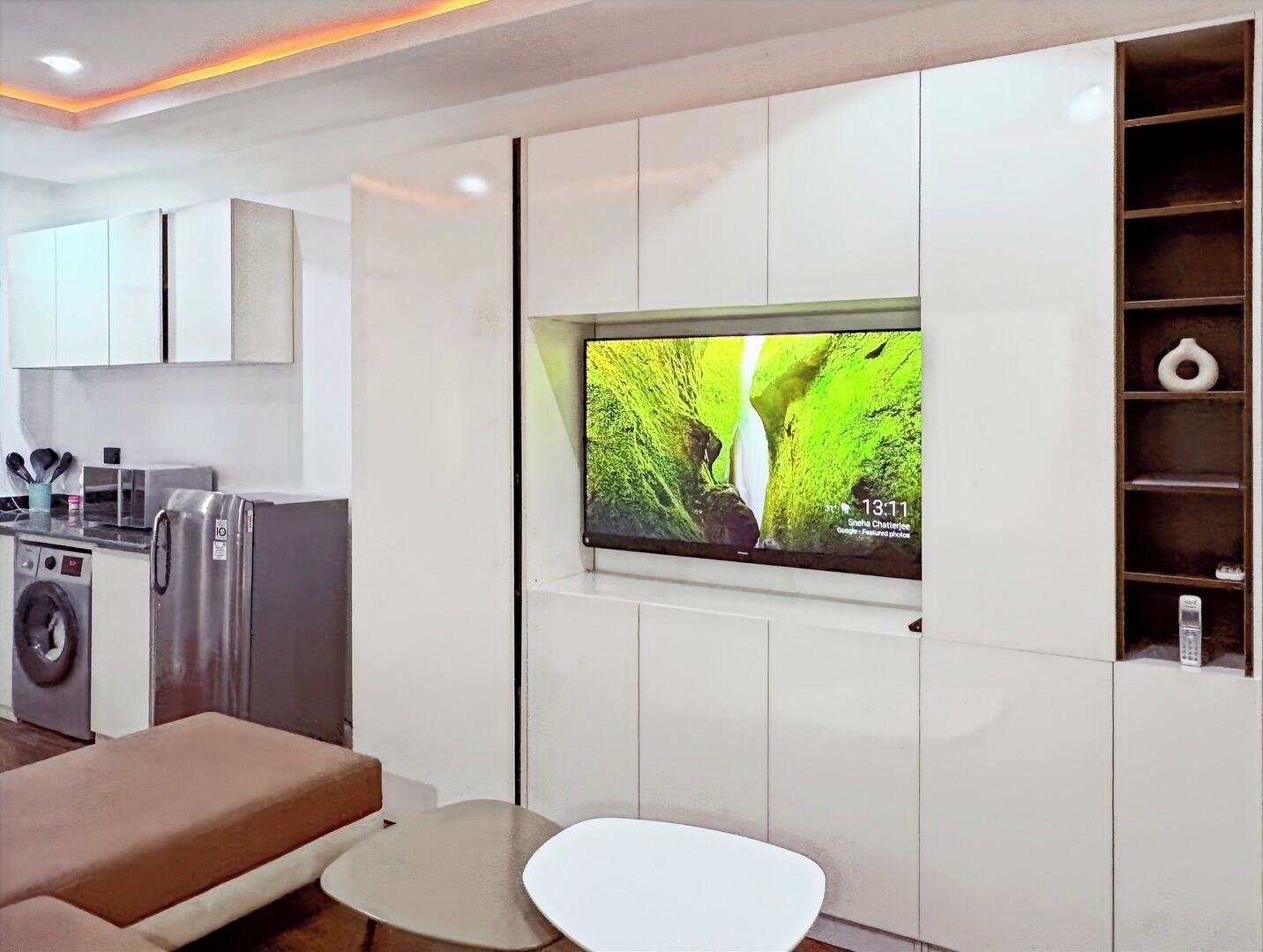 A modern living room with a flat-screen TV mounted on the wall, a sofa, a small round table, and a kitchen area with appliances.