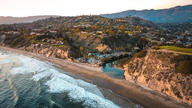 Un pueblo costero con una playa, un puente y colinas al fondo.