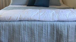 Una cama bien hecha con colcha y almohadas azul claro, dos lámparas de pared y un ventanal con cortinas.