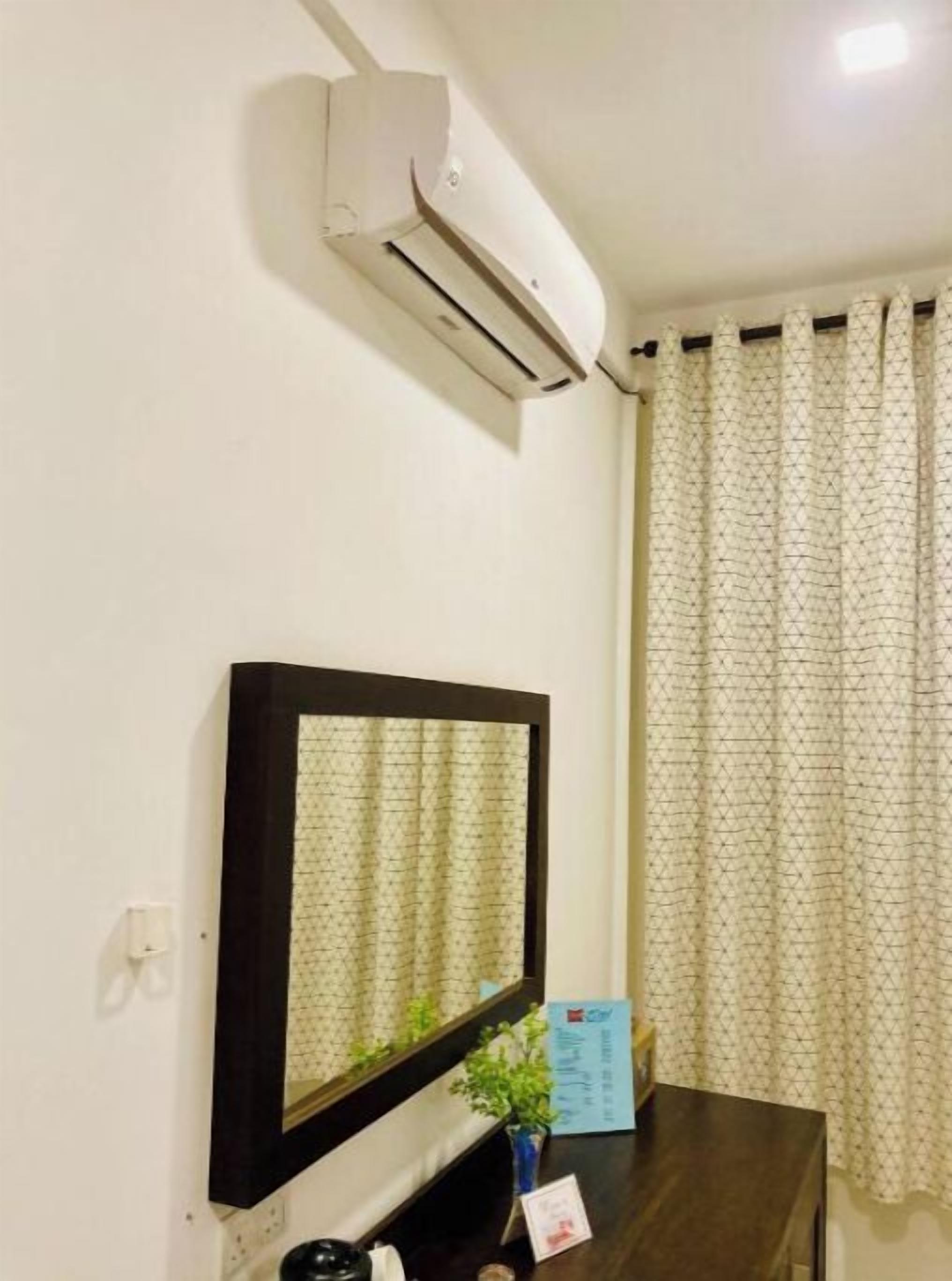 A room with a mounted air conditioner, a framed mirror, a small table with a plant, and a window with patterned curtains.