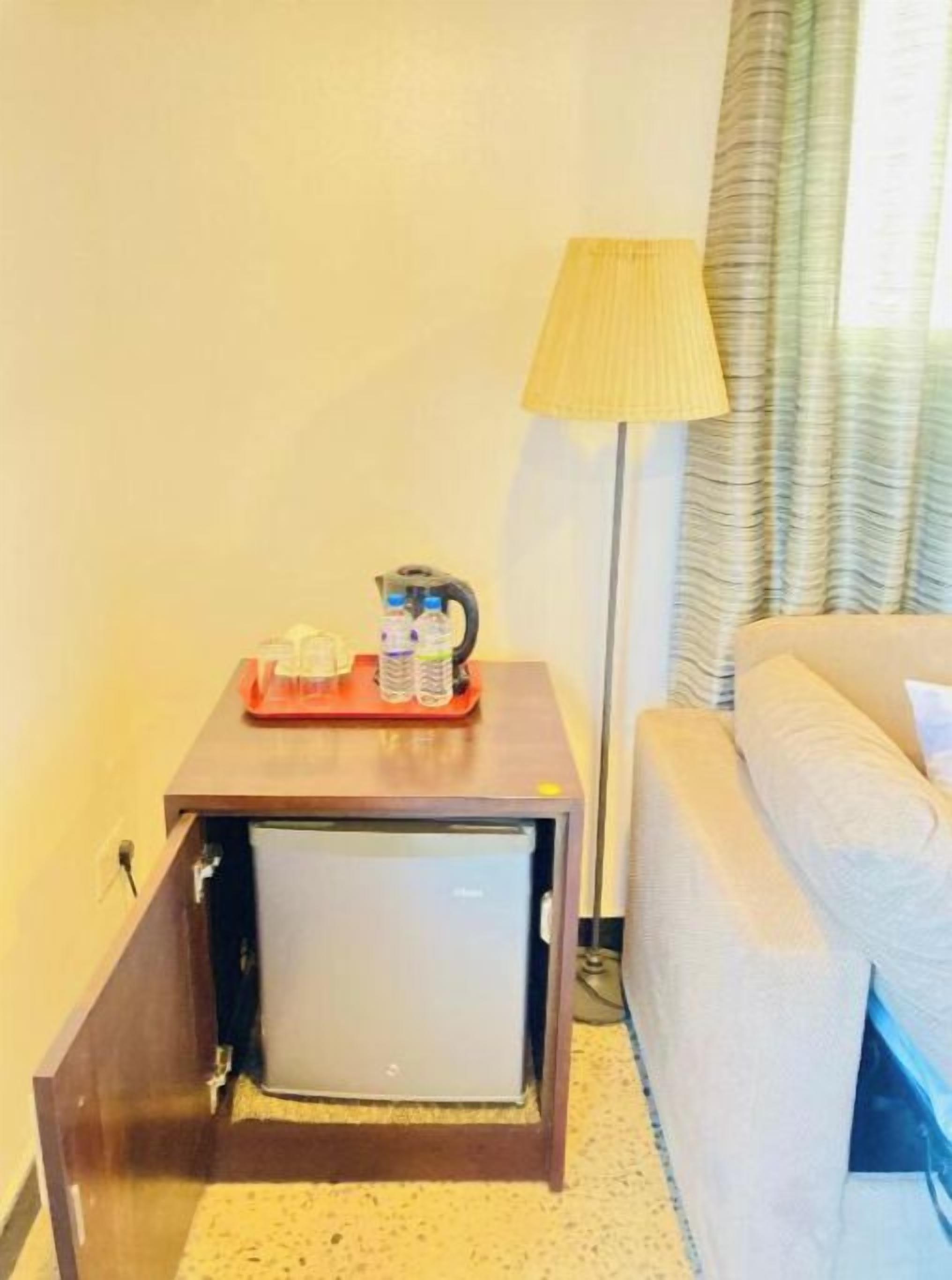 Una habitación de hotel compacta con una cama, una mesita con una lámpara y un mini bar.