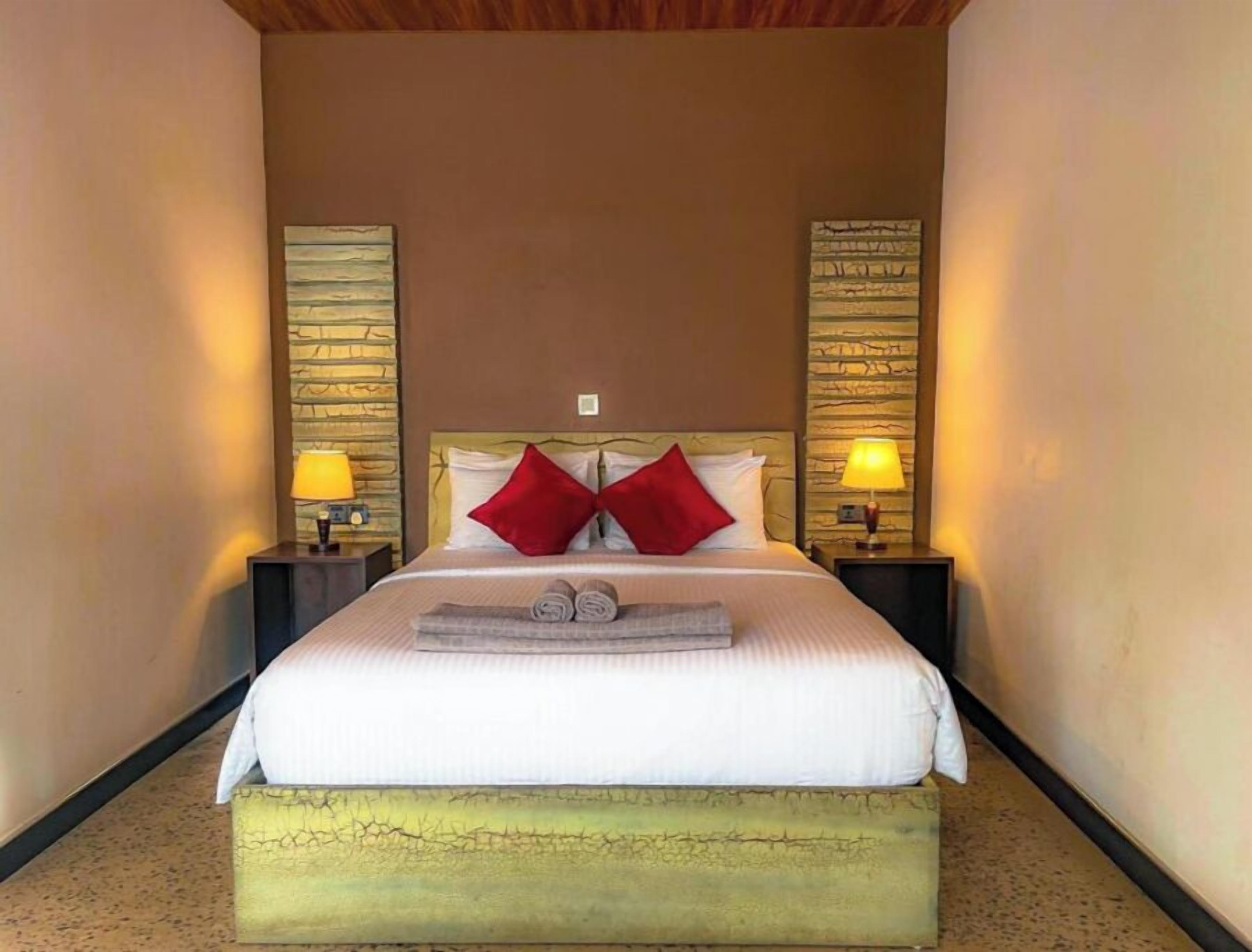 A neatly made bed with two red pillows, two bedside lamps, and a small bench at the foot of the bed.
