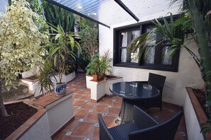 En terrasse med et rundt bord og stole, omgivet af krukker med planter og en flisegulv.