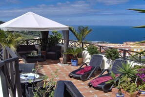 Eine Terrasse mit weißem Sonnensegel, Liegestühlen, einem Tisch mit einem Weinglas und Topfpflanzen.