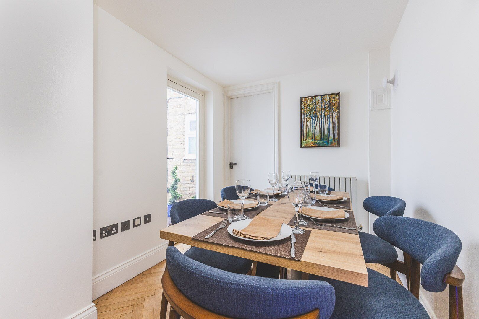 A dining area with a wooden table set for four, blue upholstered chairs, and a painting on the wall.