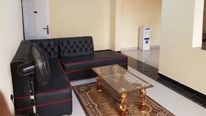 A living room with a black sectional sofa, a glass-top coffee table, and a standing fan. - Furnished Flat Apartment Bamburi Beach Mombasa (Mombasa)