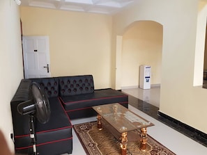 A living room with a black sectional sofa, a glass-top coffee table, and a standing fan. - Furnished Flat Apartment Bamburi Beach Mombasa (Mombasa)