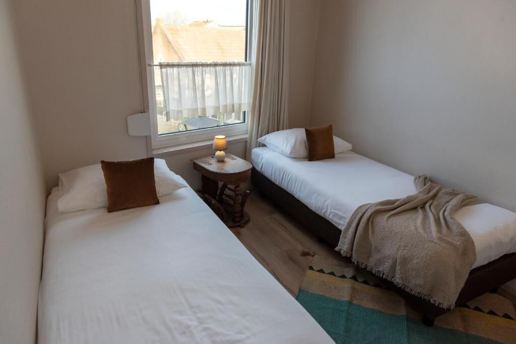 Una habitación de hotel pequeña con dos camas individuales, una mesita redonda con una lámpara y vistas al exterior a través de una ventana.